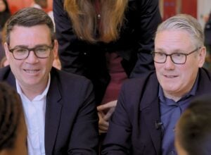 Two men wearing glasses and dark blazers sit closely together at a social or meeting setting, smiling slightly as they talk.