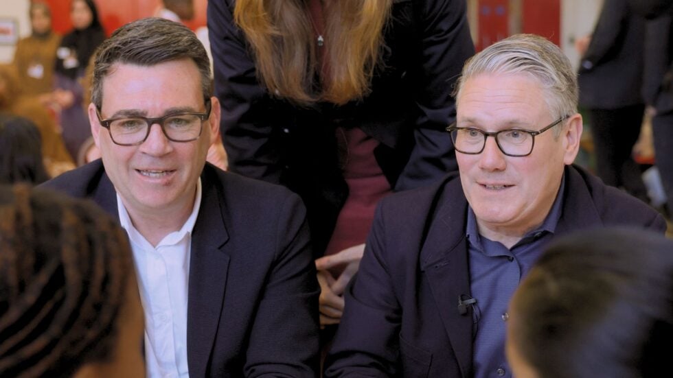 Two men wearing glasses and dark blazers sit closely together at a social or meeting setting, smiling slightly as they talk.
