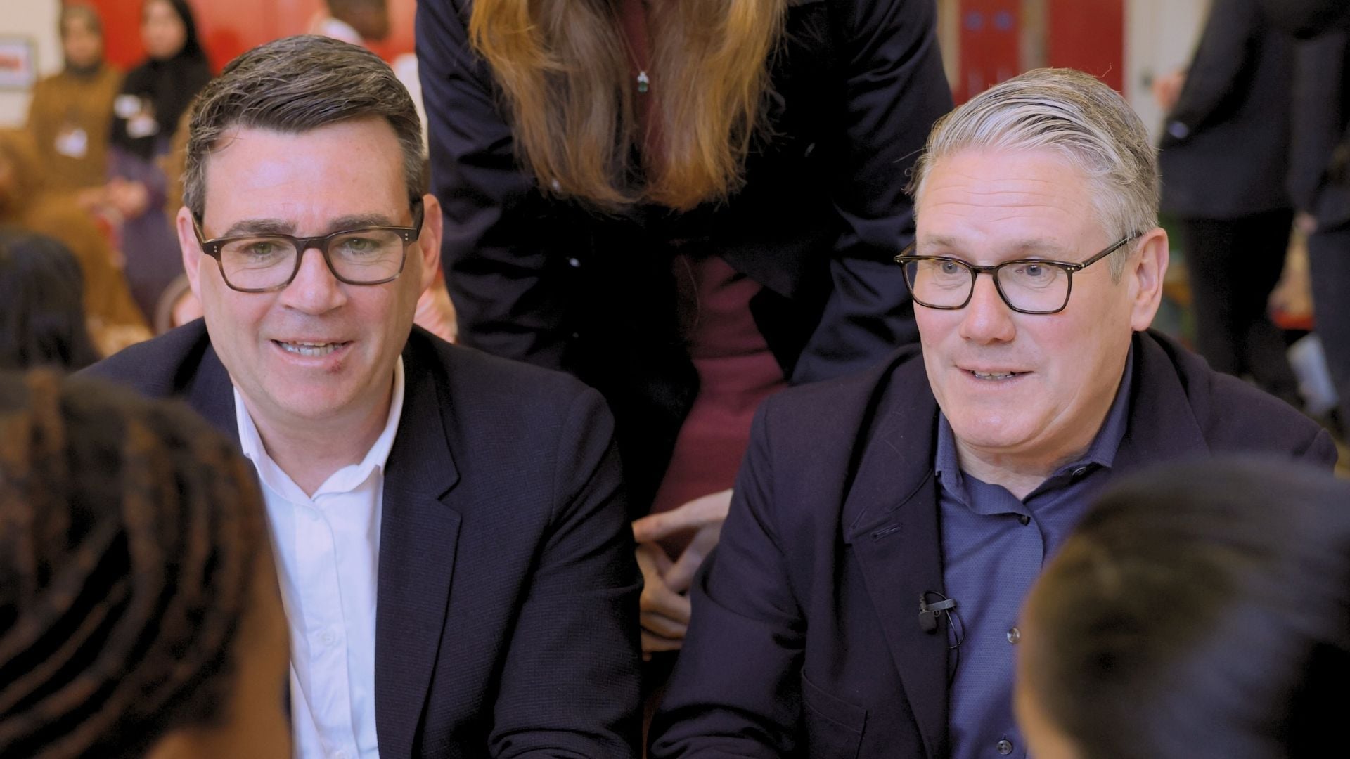 Two men wearing glasses and dark blazers sit closely together at a social or meeting setting, smiling slightly as they talk.