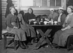 Six women wearing headscarves sit around a rough wooden table and share a meal in a simple, dim room; bowls, mugs, and a kettle visible on the table.