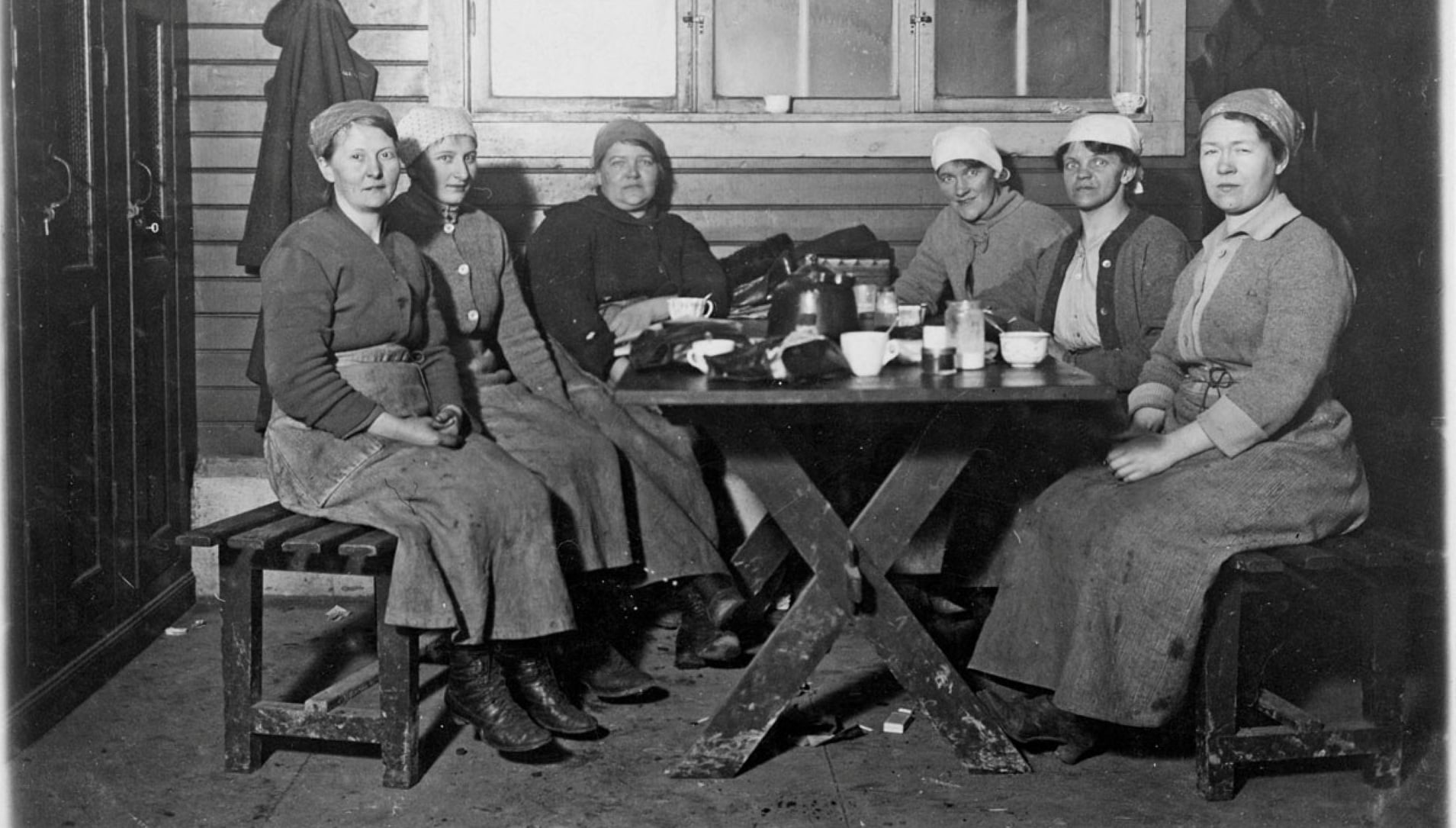 Six women wearing headscarves sit around a rough wooden table and share a meal in a simple, dim room; bowls, mugs, and a kettle visible on the table.