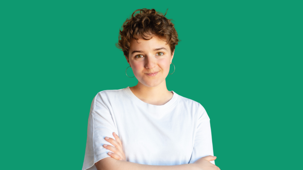 Portrait of a young woman with short curly hair, wearing a white T‑shirt, arms crossed, against a teal background.