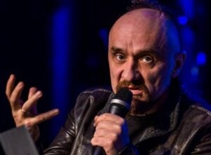 Male singer on stage holding microphone close to mouth, gesturing with his left hand under blue stage lighting