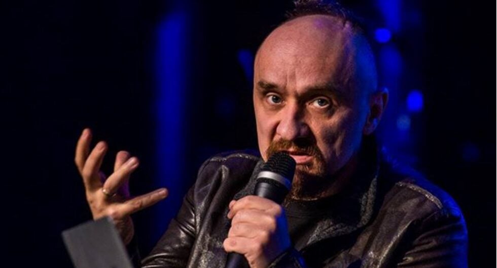 Male singer on stage holding microphone close to mouth, gesturing with his left hand under blue stage lighting