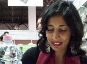 Woman with dark hair and red scarf smiling in a bookstore, shelves of books behind her and a glass globe in the foreground.