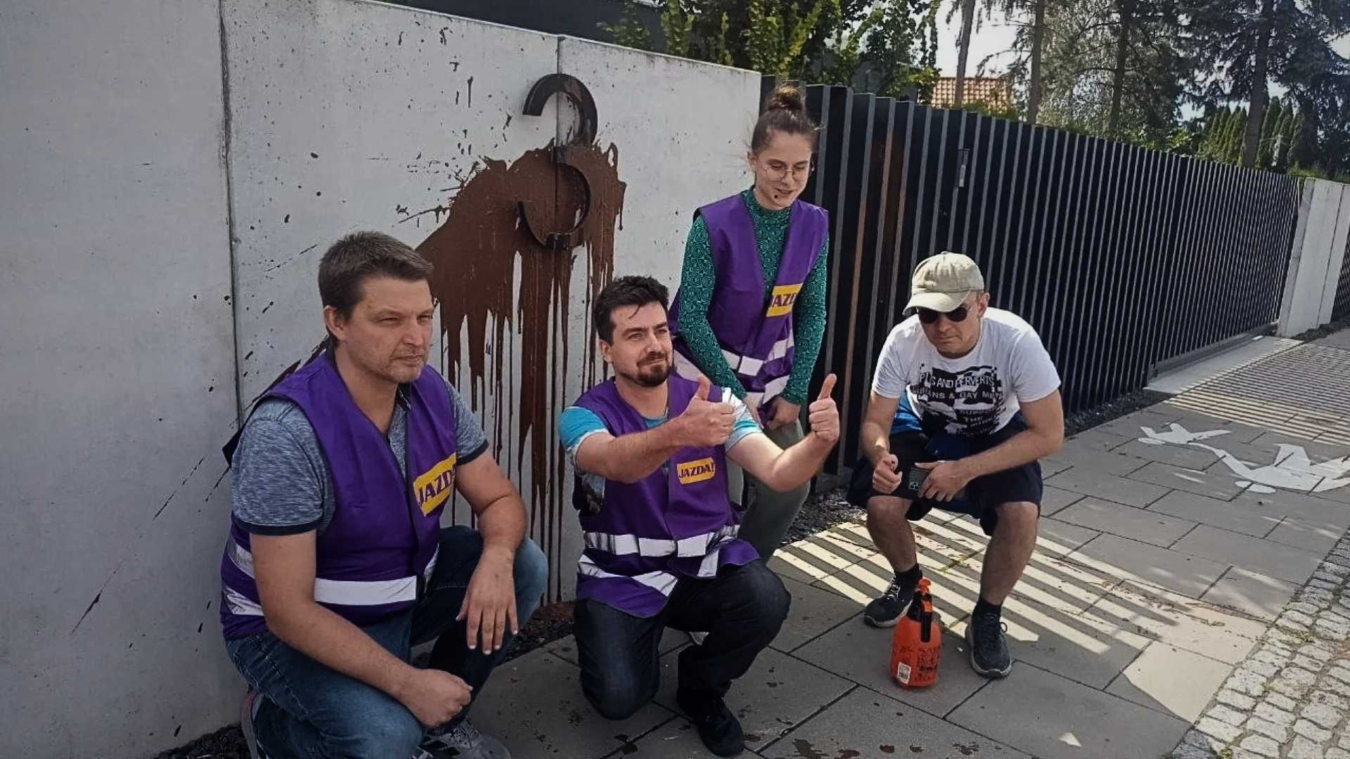 Four volunteers in purple vests pose for a photo on a sidewalk with a graffiti mural on the wall behind them, one giving a thumbs-up.