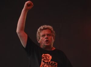 Male performer on stage with his right fist raised, under red stage lighting against a dark background.
