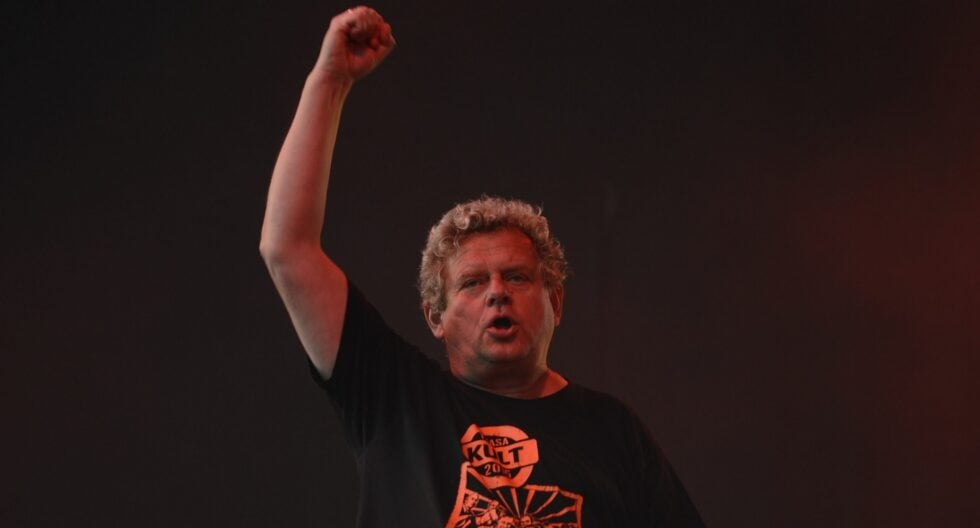 Male performer on stage with his right fist raised, under red stage lighting against a dark background.