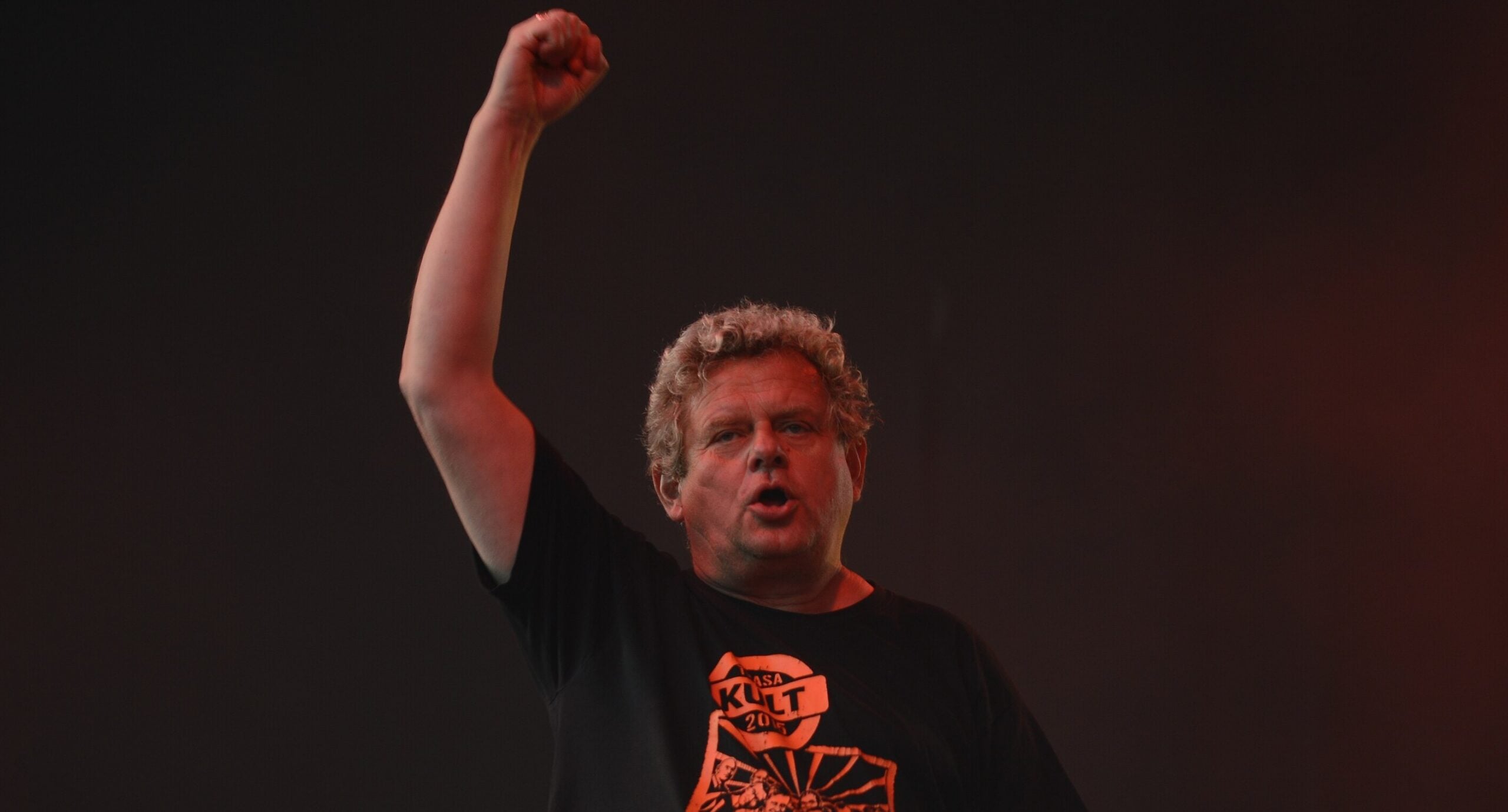 Male performer on stage with his right fist raised, under red stage lighting against a dark background.