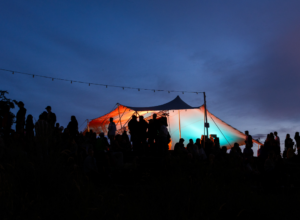 Silhouetted crowd on a hill at dusk around a large canopy tent glowing orange and teal.