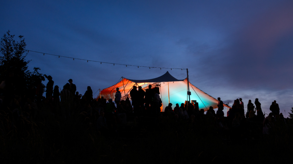 Silhouetted crowd on a hill at dusk around a large canopy tent glowing orange and teal.