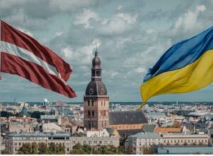 Two national flags (Latvia and Ukraine) flutter above a European city skyline, with a domed church tower in the center.