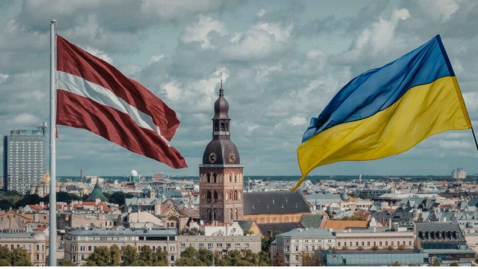 Two national flags (Latvia and Ukraine) flutter above a European city skyline, with a domed church tower in the center.