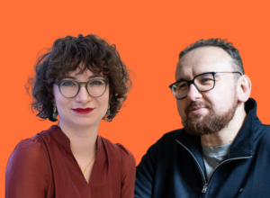 Two adults posing against an orange background: a woman with short curly hair and glasses alongside a bearded man wearing glasses.