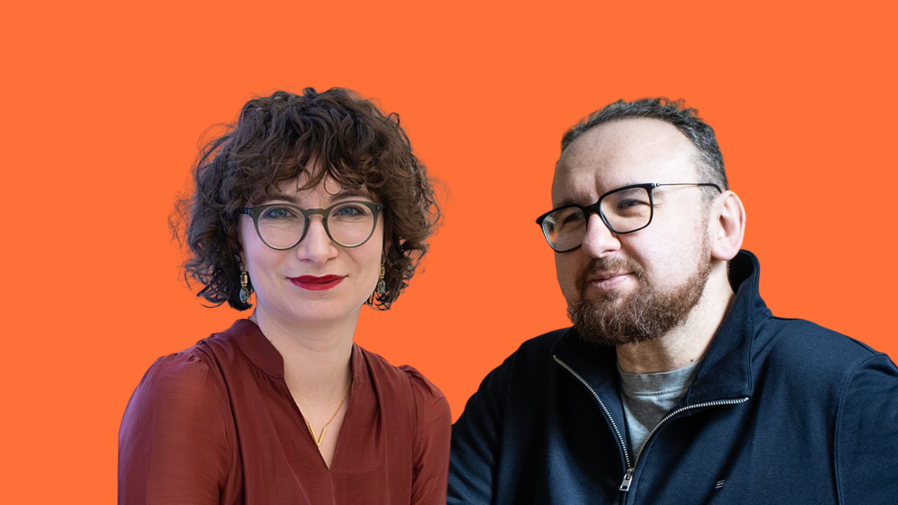 Two adults posing against an orange background: a woman with short curly hair and glasses alongside a bearded man wearing glasses.