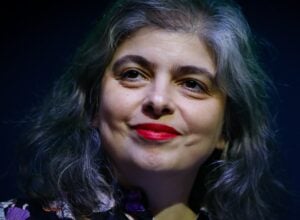 Close-up of a woman with gray hair and red lipstick smiling softly against a dark background, wearing a floral-patterned top.