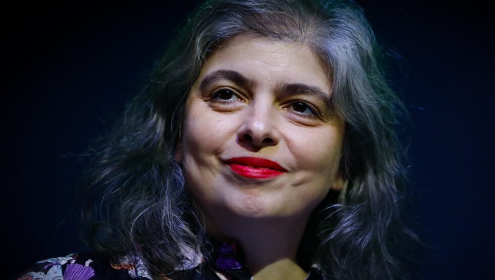 Close-up of a woman with gray hair and red lipstick smiling softly against a dark background, wearing a floral-patterned top.