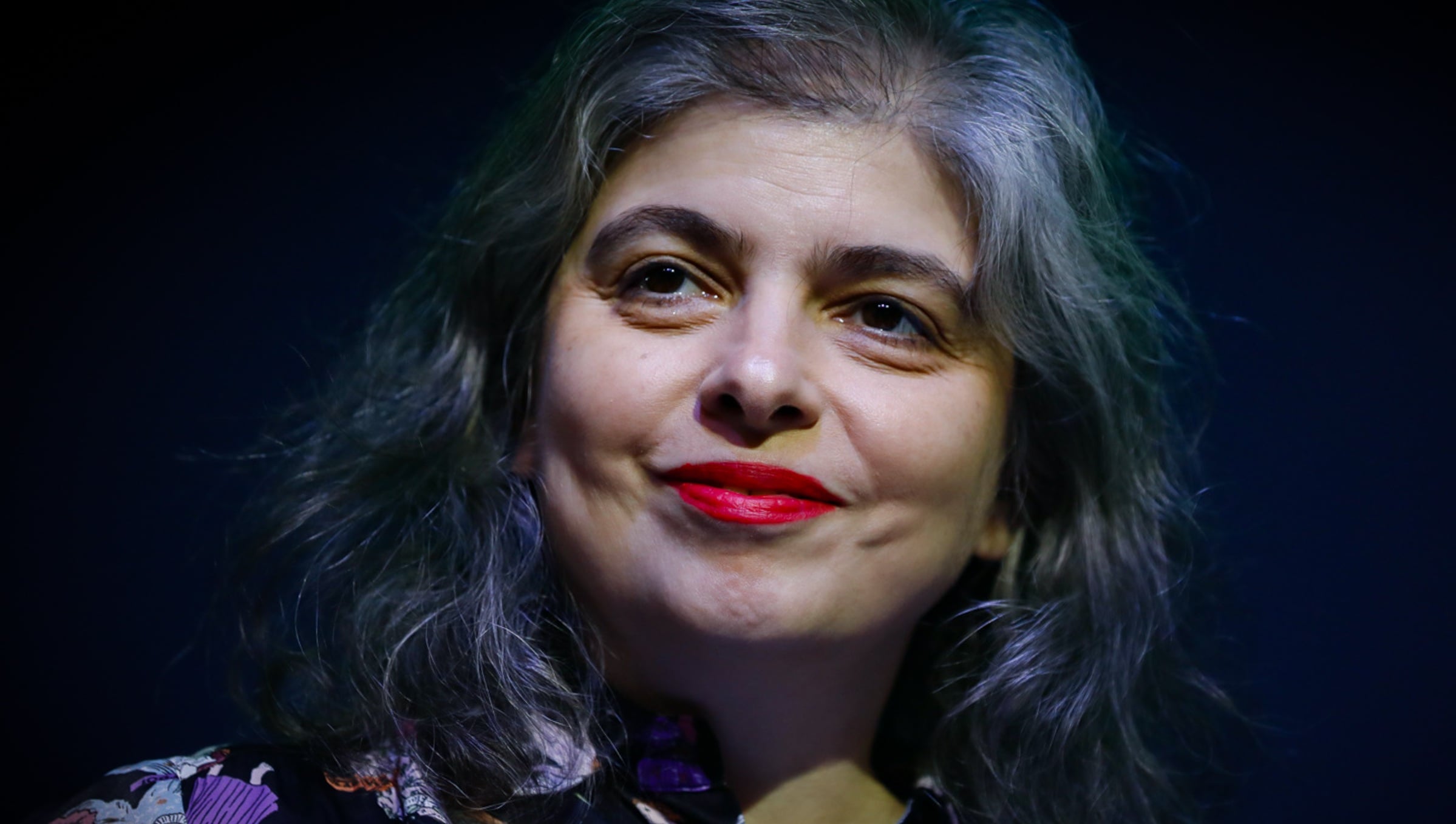 Close-up of a woman with gray hair and red lipstick smiling softly against a dark background, wearing a floral-patterned top.
