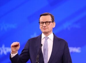 Man in a dark suit and glasses delivering a speech at a microphone against a blue backdrop.