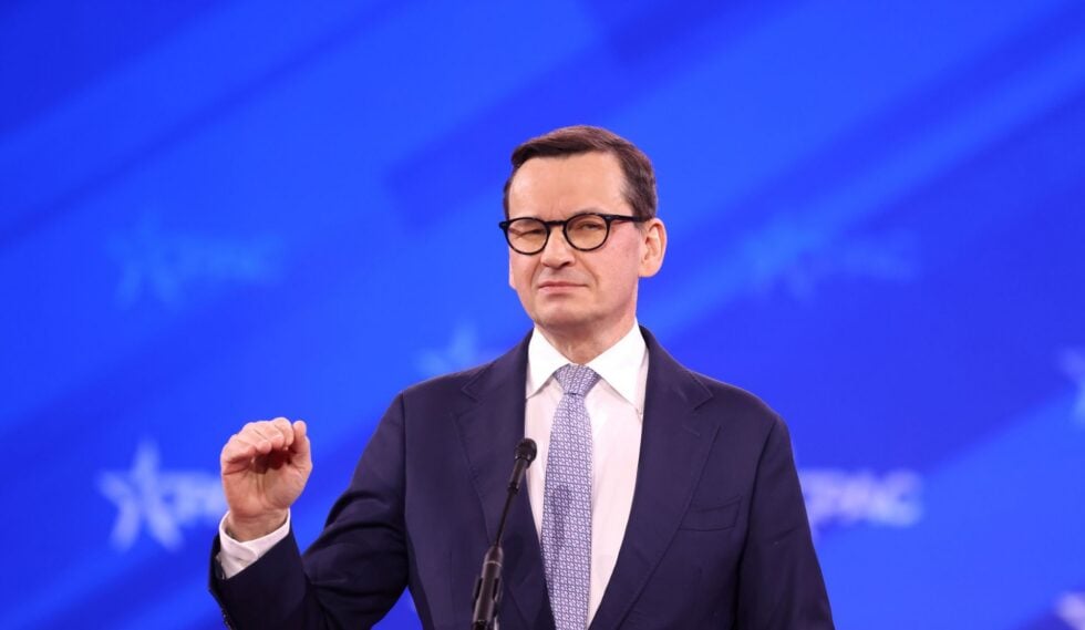 Man in a dark suit and glasses delivering a speech at a microphone against a blue backdrop.