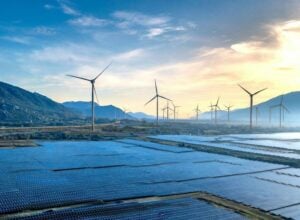 Solar panels in the foreground with wind turbines along a valley under a blue sky at sunset.