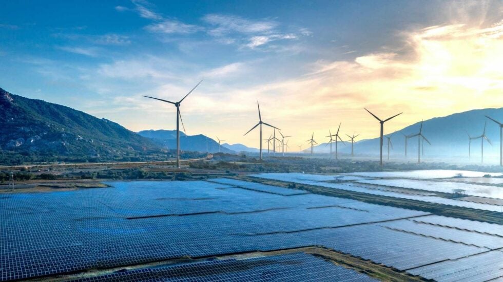 Solar panels in the foreground with wind turbines along a valley under a blue sky at sunset.