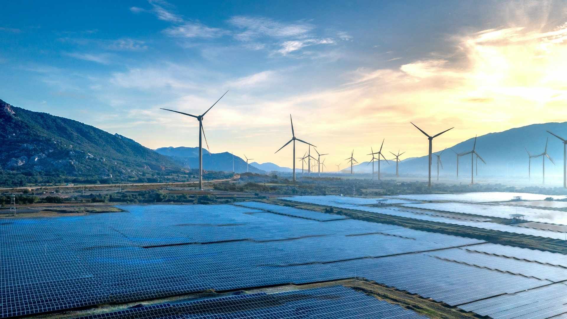 Solar panels in the foreground with wind turbines along a valley under a blue sky at sunset.