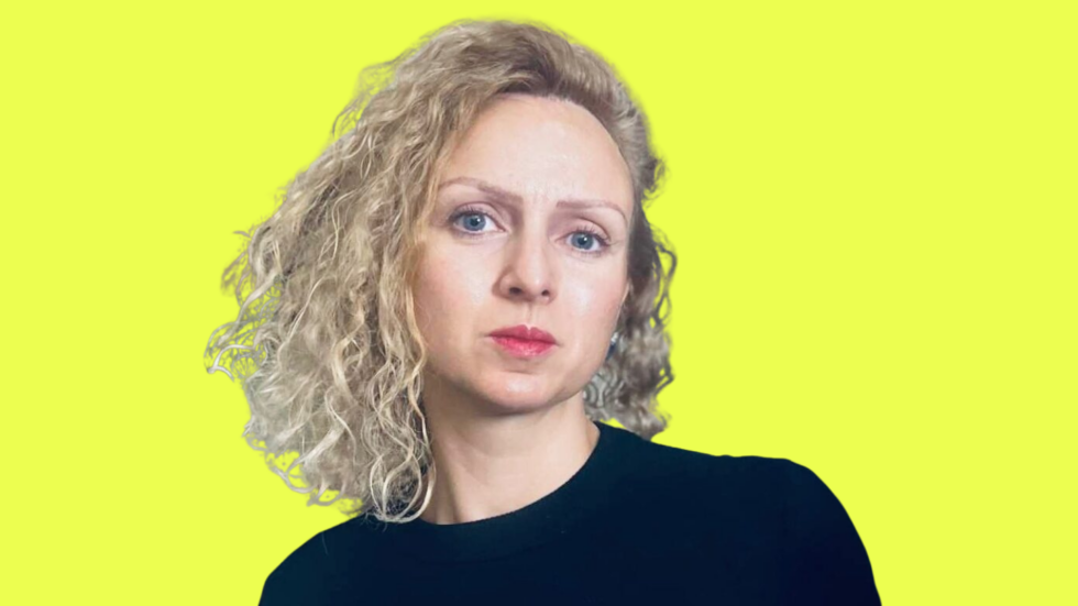 Portrait of a woman with curly blonde hair wearing a black top, looking to the right, against a bright yellow background.