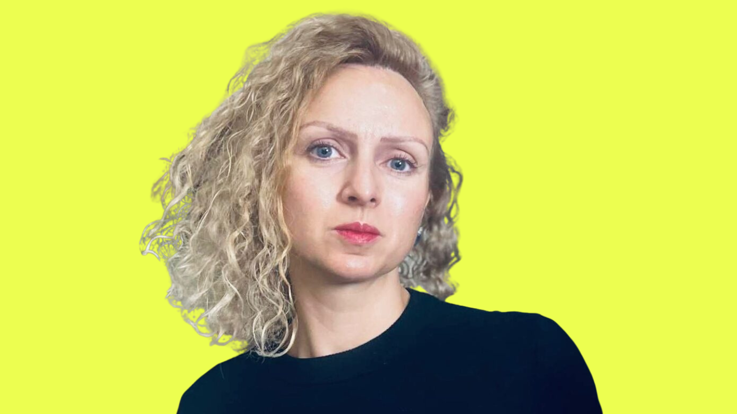 Portrait of a woman with curly blonde hair wearing a black top, looking to the right, against a bright yellow background.