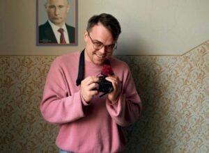 Man in a pink sweater smiles while holding a camera indoors, with vintage wallpaper and a framed portrait on the wall behind him.