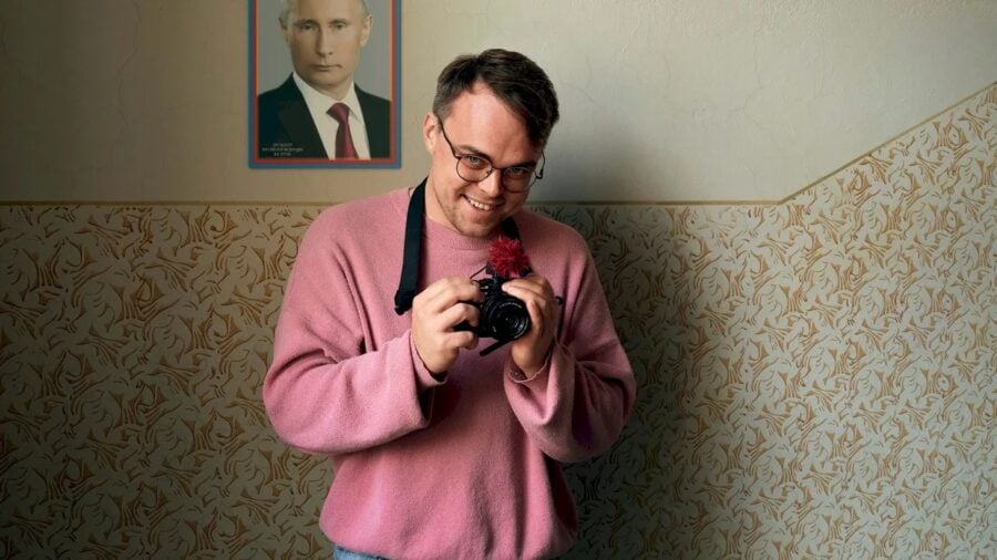 Man in a pink sweater smiles while holding a camera indoors, with vintage wallpaper and a framed portrait on the wall behind him.