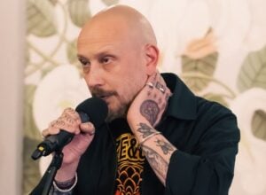 Man with a shaved head and tattoos speaks into a handheld microphone during a talk or interview.