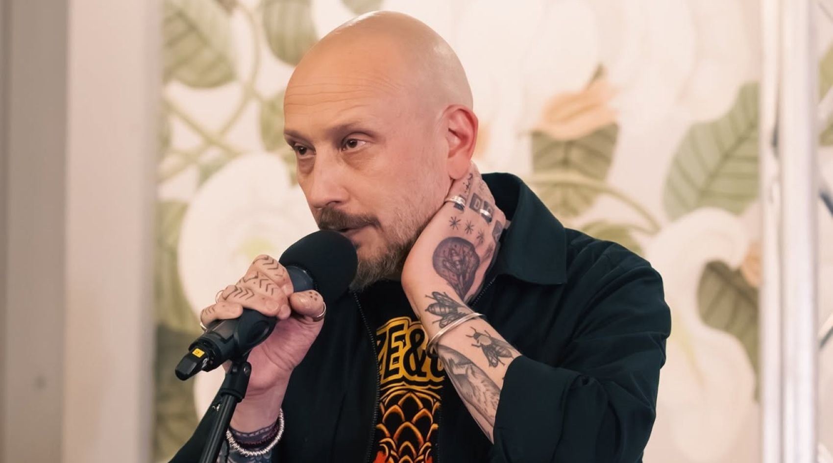 Man with a shaved head and tattoos speaks into a handheld microphone during a talk or interview.