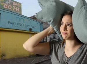 Woman shielding herself from the sun with two oversized gray pillows held over her head beside a yellow storefront and blue sign building.