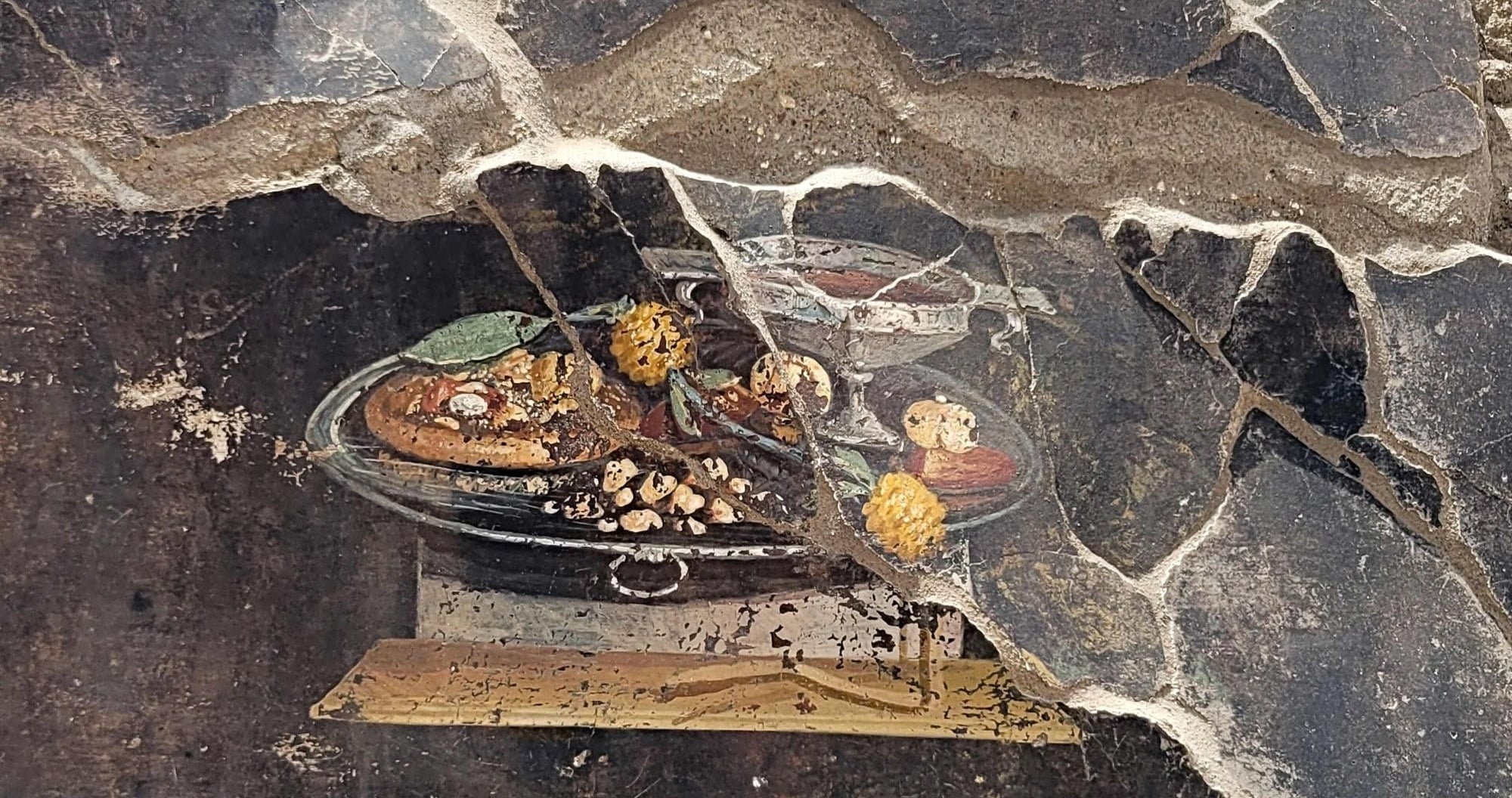 Fragment of a weathered religious painting embedded in a cracked stone wall, with dried yellow flowers nearby.