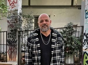 Shabtay Bendet - łysy mężczyzna w średnim wieku z siwiejącą brodą, z łańcuchem na szyi i w koszuli w biało-czarną kratę stoi uśmiechnięty na tle wysprejowanego budynku