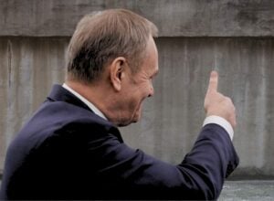 Donald Tusk widziany z prawego profilu, unosi kciuk do góry w kierunku szarego muru (fotomontaż)