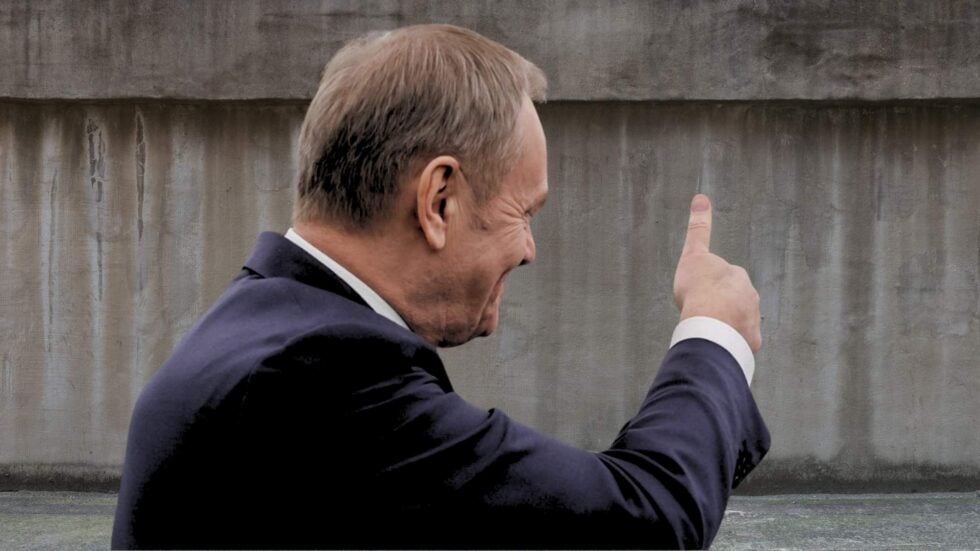 Donald Tusk widziany z prawego profilu, unosi kciuk do góry w kierunku szarego muru (fotomontaż)