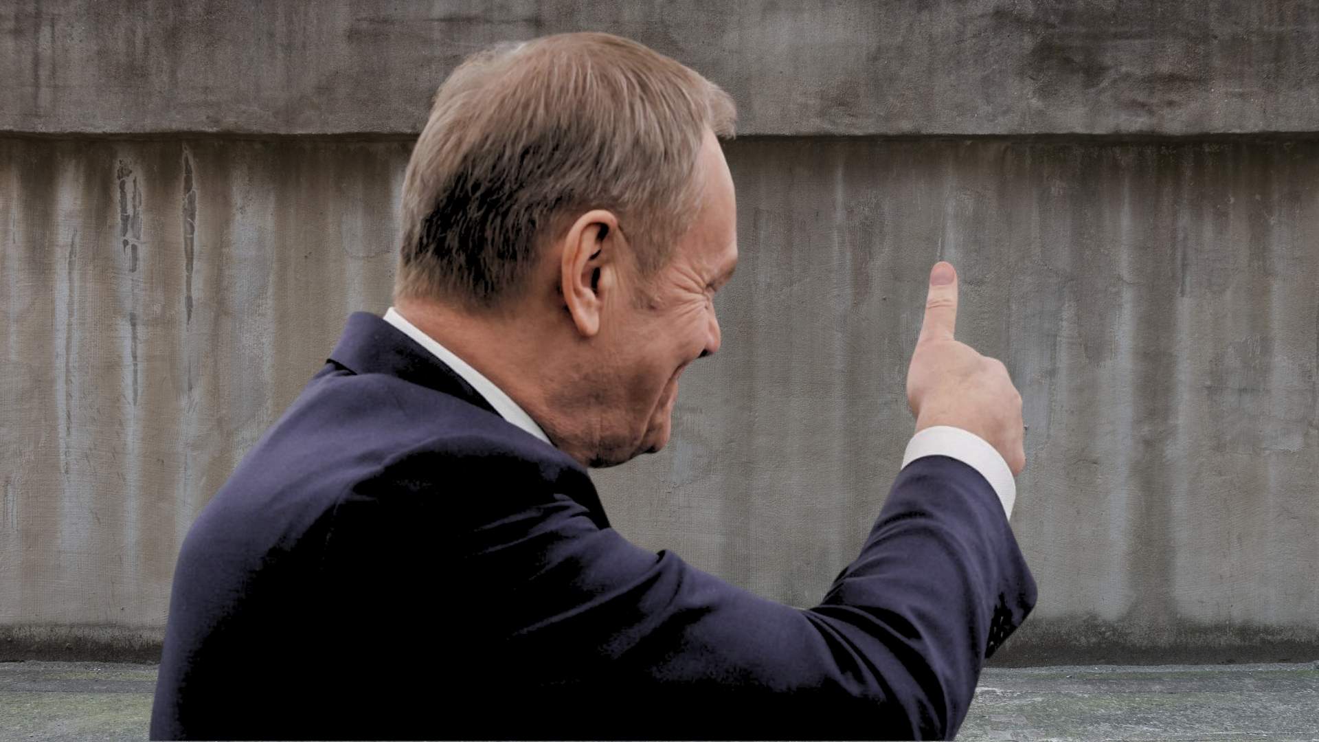 Donald Tusk widziany z prawego profilu, unosi kciuk do góry w kierunku szarego muru (fotomontaż)