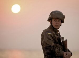 Soldier in camouflage gear stands at sunset, with Ukrainian flag patch on the sleeve, looking to the side.