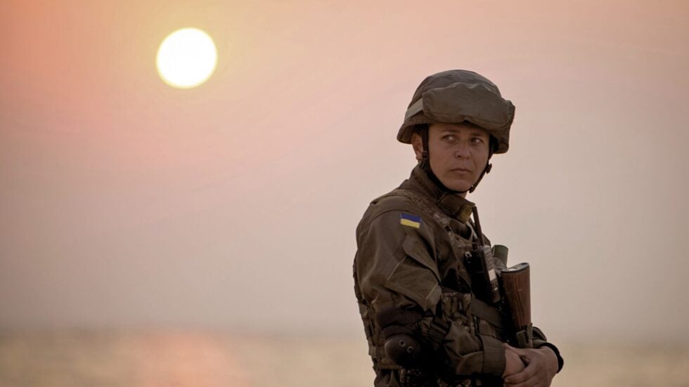 Soldier in camouflage gear stands at sunset, with Ukrainian flag patch on the sleeve, looking to the side.