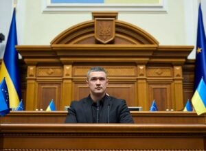 Male official in a black suit speaks from a wooden podium in a grand chamber with Ukrainian and EU flags nearby.
