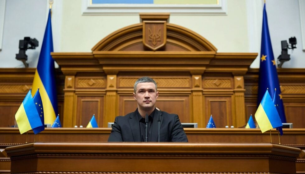 Male official in a black suit speaks from a wooden podium in a grand chamber with Ukrainian and EU flags nearby.