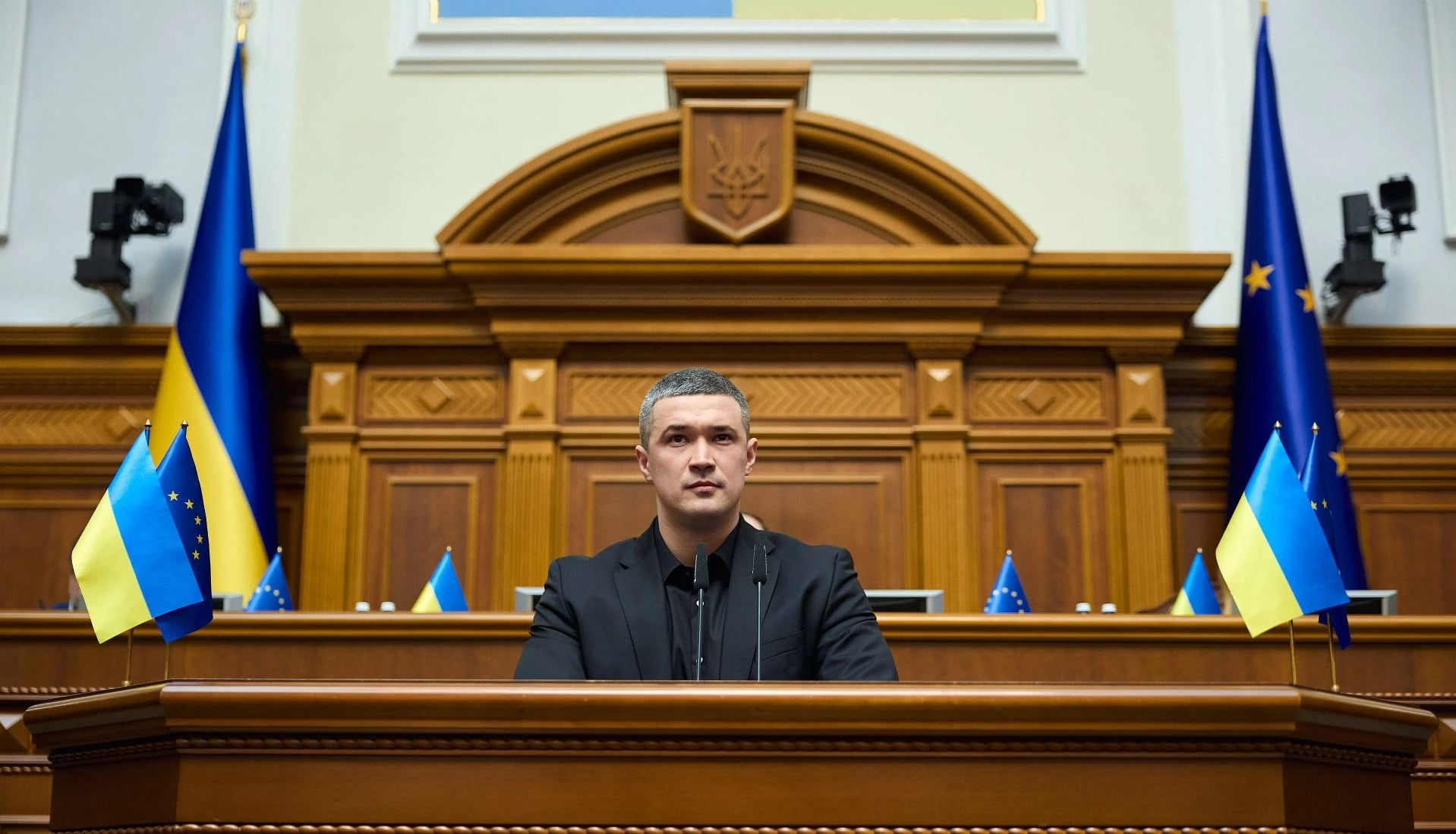 Male official in a black suit speaks from a wooden podium in a grand chamber with Ukrainian and EU flags nearby.