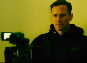 Man in a dark hoodie stands in a yellow-lit room, facing a video camera in the foreground, serious expression.