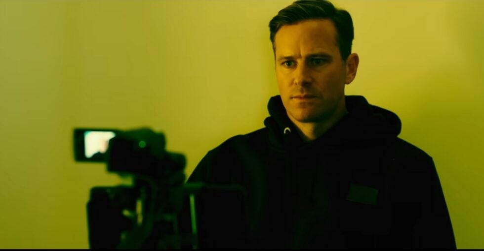 Man in a dark hoodie stands in a yellow-lit room, facing a video camera in the foreground, serious expression.