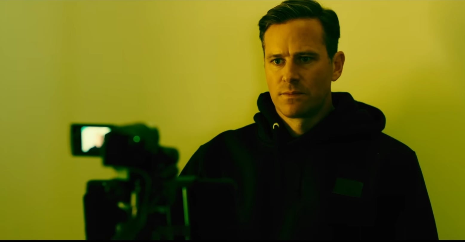Man in a dark hoodie stands in a yellow-lit room, facing a video camera in the foreground, serious expression.