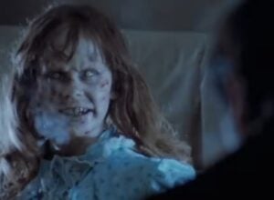 Pale zombie woman with a blood-streaked face and ragged hair in a tattered blue dress, in a dark scene.