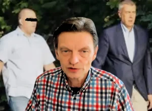 Presenter in a red-blue plaid shirt speaks to camera with two blurred men in a forest-like background behind him