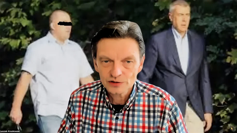 Presenter in a red-blue plaid shirt speaks to camera with two blurred men in a forest-like background behind him
